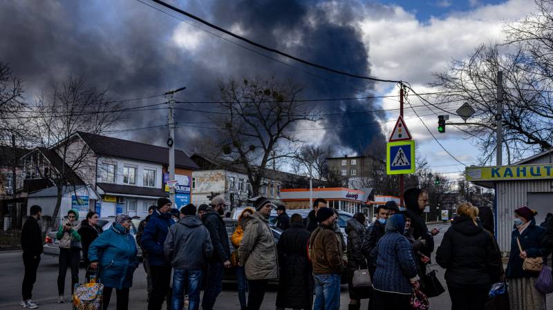 People stand in line in front of a supermarket while smoke billows over the town of Vasylkiv just outside Kyiv after overnight Russian strikes hit an oil depot. AFP